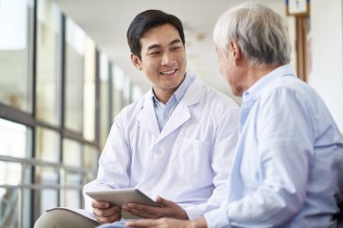 Konuşan ve test sonuçlarını hastanedeki yaşlı hastaya açıklayan gülümseyen Asyalı genç doktor.