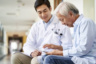 Konuşan ve test sonuçlarını hastanedeki yaşlı hastaya açıklayan gülümseyen Asyalı genç doktor.