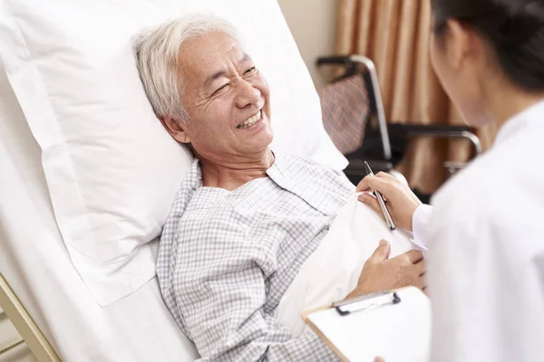 asian old man lying in bed talking to young female doctor in hospital ward