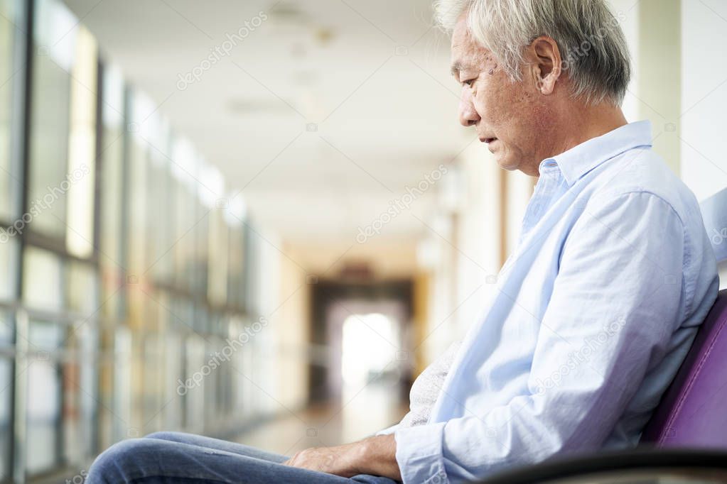 sad and devastated asian senior man sitting alone in empty hospital hallway, head down