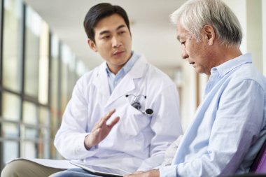 Genç Asyalı doktor konuşuyor ve hastane koridorunda morali bozuk yaşlı hastaya test sonuçlarını ve teşhisi açıklıyor.