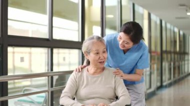 Dost canlısı Asyalı işçi, destekli yaşam merkezinin koridorunda tekerlekli sandalyeye bağlı bir ihtiyarla konuşuyor.