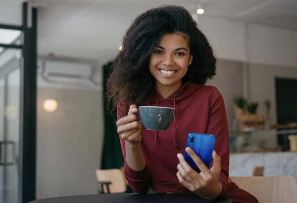 Happy beautiful woman drinking coffee, holding mobile phone, communication in modern cafe. Coffee break. Young emotional African American hipster using  application for smartphone, looking at camera