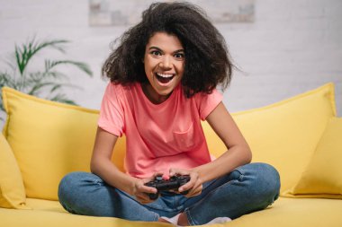 Mutlu duygusal kadın video oyunları oynuyor, sarı koltukta oturuyor, elinde kablosuz joystick var. Heyecanlı Afrikalı Amerikalı hippi kız eğleniyor, evde kalıyor.  