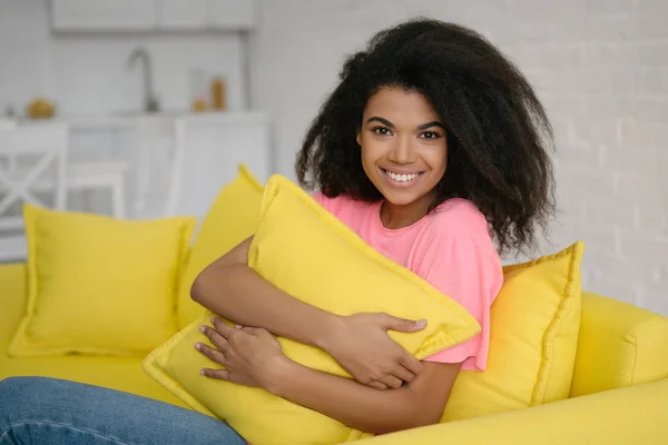 年轻美丽的非洲裔美国女人抱着枕头坐在客厅舒适的黄色沙发上 可爱的 情绪化的卷发女孩在家里放松和微笑的真实写照 — 图库照片