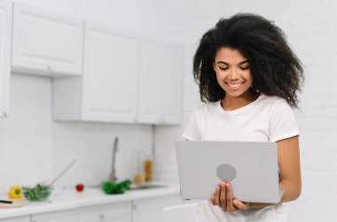 Mutfakta durmuş yemek kurslarını izleyen gülümseyen genç bir kadın. Mutlu Afro-Amerikan kadın dizüstü bilgisayar ve internet kullanıyor, evde serbest çalışıyor.