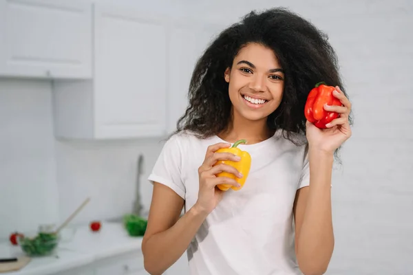 Beautiful African American woman choosing between two bell peppers standing in the kitchen. Healthy lifestyle concept, vegetarian, diet. Happy housewife holding organic vegetables, cooking fresh salad at home