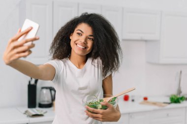 Güzel Afro-Amerikan yemek blogcusu mutfakta vejetaryen yemeği pişiriyor, internetten video yayınlıyor. Sağlıklı yaşam tarzı kavramı. Mutlu duygusal kadın taze salatayla bağırıyor, selfie çekiyor.