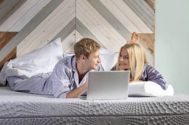 Yatak odasındaki genç adam ve kadın bilgisayara bakar, kulaklıkla müzik dinler.