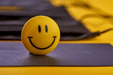 Siyah yüzeyde duran kameraya gülen bir yüz bakıyor. Arkasında sarı kağıtta siyah maskeler var. Renkli. Karantinaya alındı. Sağlık en önemli şeydir..