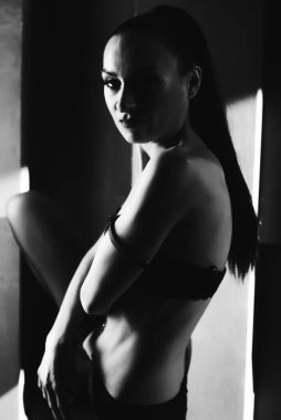 Seksi kız modelini duvarın arka planına karşı güçlendiriyor. Güneş ışığı. Siyah moda keten giymiş ince bir kız. Kameraya bakıyor. BW. Göğüs bandını indirdim. yan görünüm.