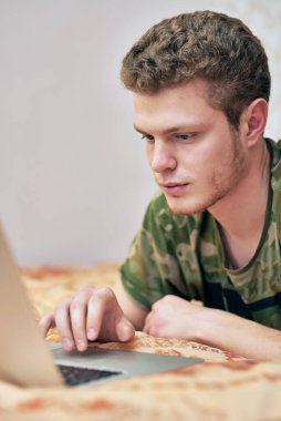 Yeşil tişörtlü adam karantinada, evin kanepesinde yatıyor ve dokunmatik pede basan dizüstü bilgisayara bakıyor. Evde bilgisayar başında eğlenir, çalışır ve ders çalışırdı. Opetercovid-19. Evde kal.