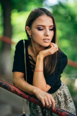 Güzel esmer kız yan yana bakar, ellerini tırabzana yaslar, boruya yaslar. Güzel kahverengi gözler, siyah ceket. Yeşil doğa. seksi ledy.