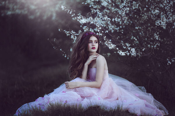 Beautiful Romantic Girl with long hair in fairy long pink dress sitting near flowering tree.