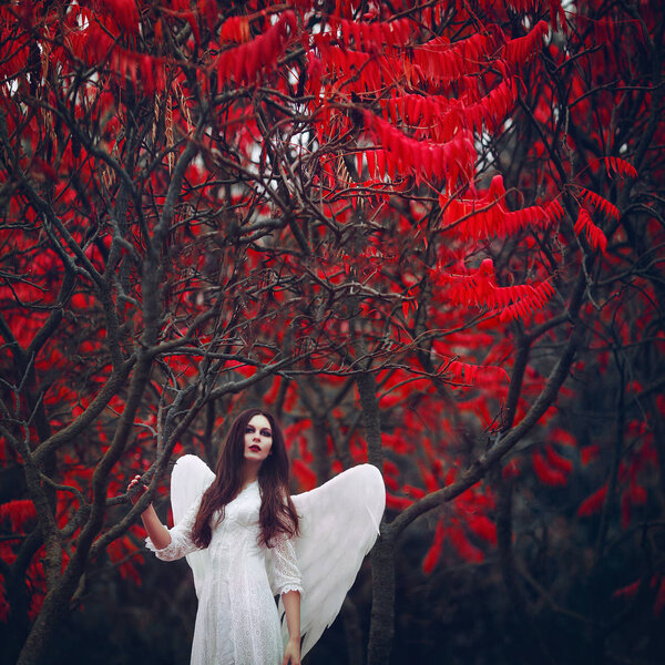 Art photo of a Angelic beautiful woman. A girl with angel wings and a white dress near blood-red trees.