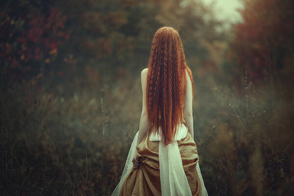 A beautiful young woman with very long red hair as a witch walks through the autumn forest. Back view. Creative colors and Artistic processing.