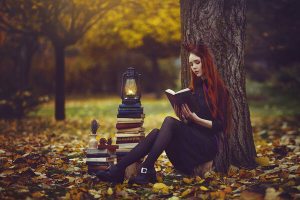 Beautiful red-haired girl with books and a lantern sitting under a tree in the autumn fairy forest. A fabulous autumn photo shoot.