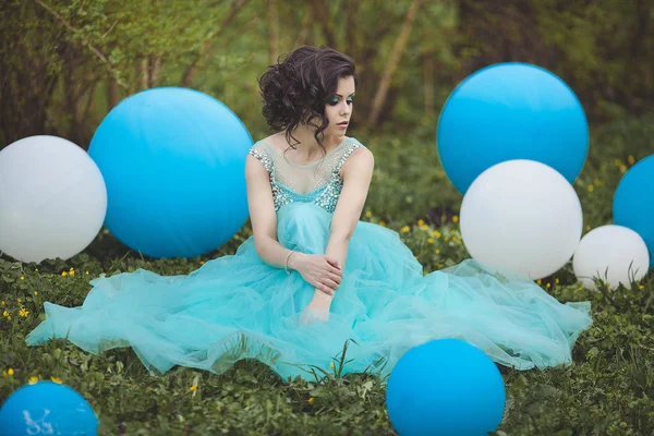 Beautiful girl graduate in a blue dress is sitting on the grass near a large blue and white balloons. Pensive elegant young woman in a beautiful dress.