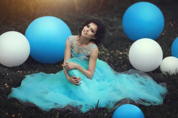 Happy girl in prom with helium air balloons..Beautiful girl graduate in a blue dress is sitting on the grass near a large blue and white balloons. Lovely girl with a beautiful smile.