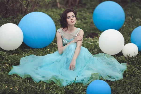在舞会上带着氦气球的快乐女孩。一个穿着蓝色连衣裙的漂亮女孩坐在草地上, 旁边是一个大的蓝色和白色的气球。开朗优雅的年轻女子穿着漂亮的礼服在 — 图库照片