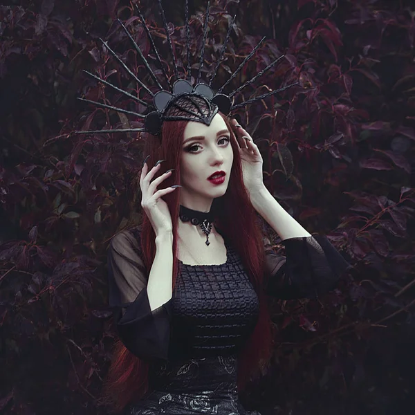 Una bella donna con la pelle chiara e lunghi capelli rossi in un abito nero e nel crownk nero. Strega ragazza con artigli di vampiro e labbra rosse. Aspetto gotico. Vestito per Halloween . — Foto Stock