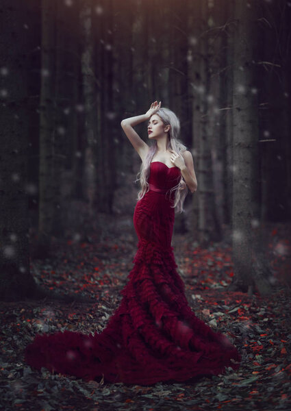 Beautiful woman with long white hair posing in a luxurious red dress with a long train standing in a autumn pine forest. Creative colors and Artistic processing.