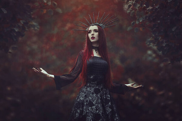 Красивая женщина с бледной кожей и длинными рыжими волосами в черном платье и в черной короне. Ведьма с вампирскими когтями и красными губами. Готический вид. Наряд на Хэллоуин
.
