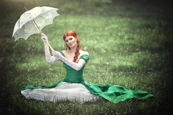 Beautiful young red-haired girl in a medieval green dress with an umbrella sitting on the grass. — Stock Photo, Image