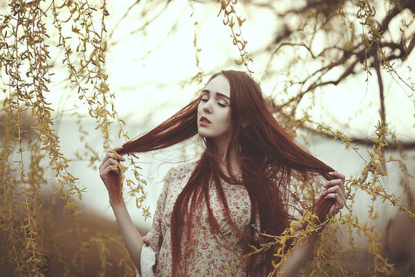 Portrait of a romantic girl with red long hair in the wind under a willow tree.