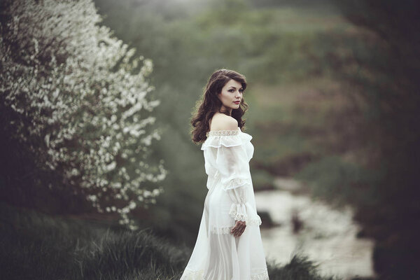 On the edge of the shore near the river there is a girl in a white dress near a flowering tree. The girl in the wind in the spring. Longing for freedom concept
