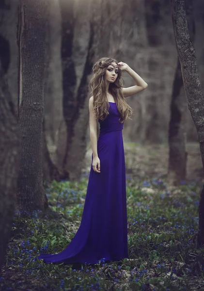 Elfo ragazza con i capelli lunghi e gli occhi azzurri in tiara e un abito lungo blu con un treno a piedi attraverso la foresta in fiore primavera. Ragazza principessa sogni . — Foto Stock