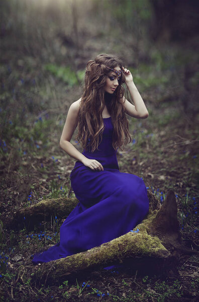 Elf girl with long hair and blue eyes in the tiara and a long blue dress sitting on a stump in the spring . Girl Princess dreams. Creative colors and Artistic processing.