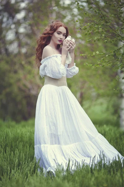 Una sensual chica pelirroja con un vestido blanco sostiene una paloma blanca en su mano. La paloma blanca es un símbolo de paz . —  Fotos de Stock