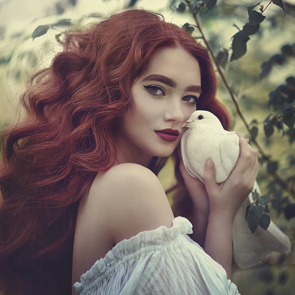 Una sensuale ragazza dai capelli rossi in un abito bianco con le spalle nude tiene in mano una colomba bianca. La colomba bianca è un simbolo di pace . — Foto Stock