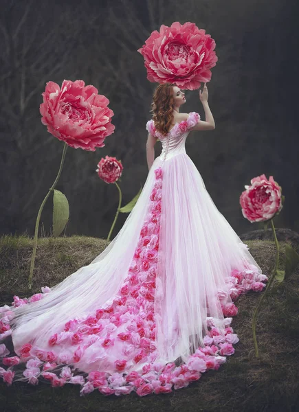 Menina sensual bonita com cabelo vermelho em um vestido rosa fada com um trem enorme posando perto das flores gigantes de uma peônia rosa. A menina é uma princesa flor . — Fotografia de Stock