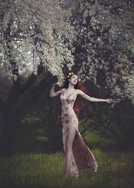 Mooie jonge vrouw met zeer lange rode haren in een gouden jurk onder een bloeiende boom, wandelen door het bos van de lente. Lange rode haren ontwikkelt in de wind. — Stockfoto