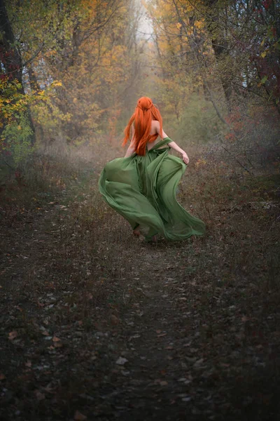 Een langharig roodharig elfenmeisje loopt de verte in door het mistige herfstbos. Een feeënvrouw met lang haar in een middeleeuwse jurk. — Stockfoto