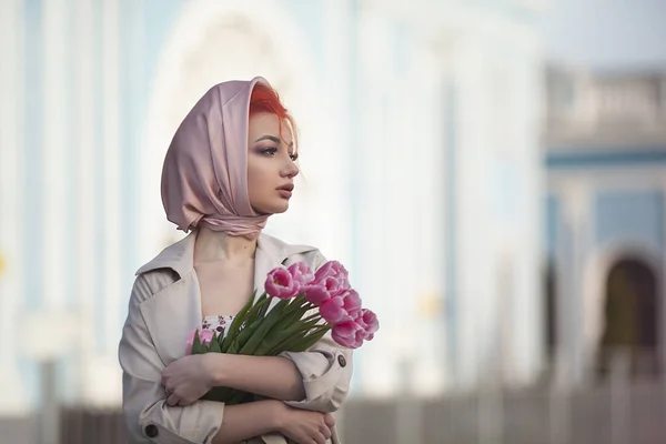 Een vrouw in een lente jurk en jas, als een oude stijl, houdt een boeket van roze bloemen in de oude stad. Romantisch retro meisje loopt rond in de stad. — Stockfoto