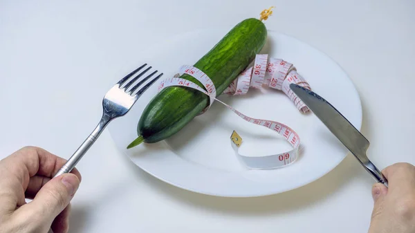 Az étrend és az egészséges életmód fogalma. A férfi kezek felülnézete egy asztalnál, villával és késsel egy lapos fehér tányéron, amiben egy mérőszalaggal összekötött uborka fekszik.. — Stock Fotó