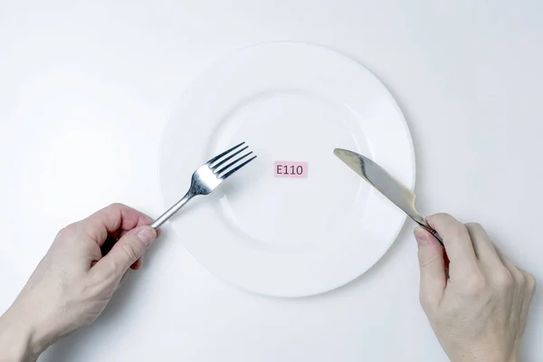 Káros élelmiszer-adalékanyagok. A férfiak kezei kést és villát tartanak. A tányéron van egy E-additív kóddal ellátott lemez.. — Stock Fotó