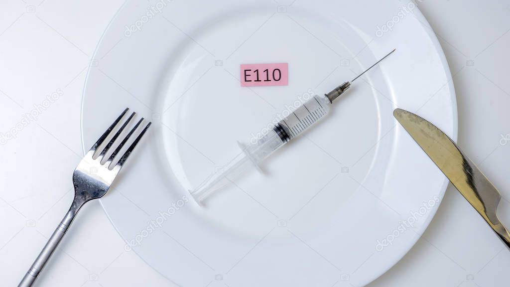 Harmful food additives. Knives, fork, plate. On the plate is a syringe and an E-supplement code plate.