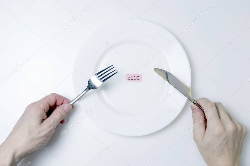 Harmful food additives. Men's hands hold a knife and a fork. On the plate is a plate with the E-additive code.
