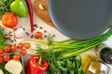 Kesme tahtasının üzerinde siyah bir tabak duruyor ve sebzelerin etrafında yeşiller var.