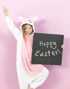 pretty blonde girl in cozy rabbit costume holding white board with text happy easter and posing in studio 