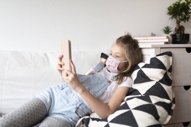 Korunaklı maskeli genç kız Corona Covid 19 krizi karantinası sırasında evde tablet bilgisayar kullanıyor.