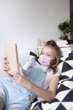 Korunaklı maskeli genç kız Corona Covid 19 krizi karantinası sırasında evde tablet bilgisayar kullanıyor.