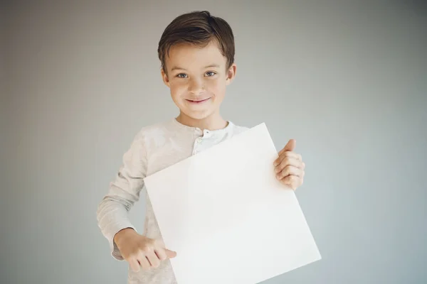 Крутой Мальчик Держащий Белый Пустой Лист Перед Серым Фоном — стоковое фото