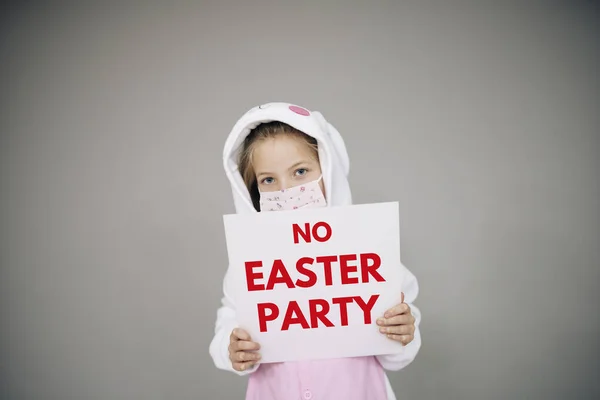 beautiful girl in easter bunny costume and face protective mask holding white board sign with text no easter party during corona lockdown