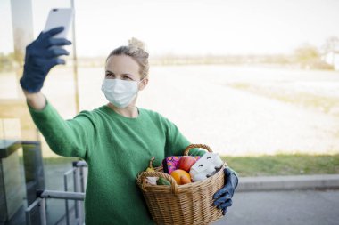 Yüzü maskeli, yeşil kazaklı ve siyah eldivenli güzel bir kadın alışveriş yaptıktan sonra bir telefon görüşmesi yapıyor ve alışveriş sepetini tutuyor.