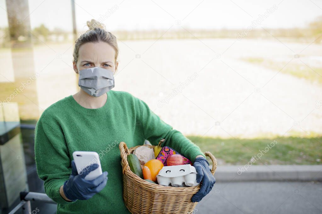 Pretty woman with face mask and green pullover and black gloves makes a phone call after shopping and is holding her shopping basket